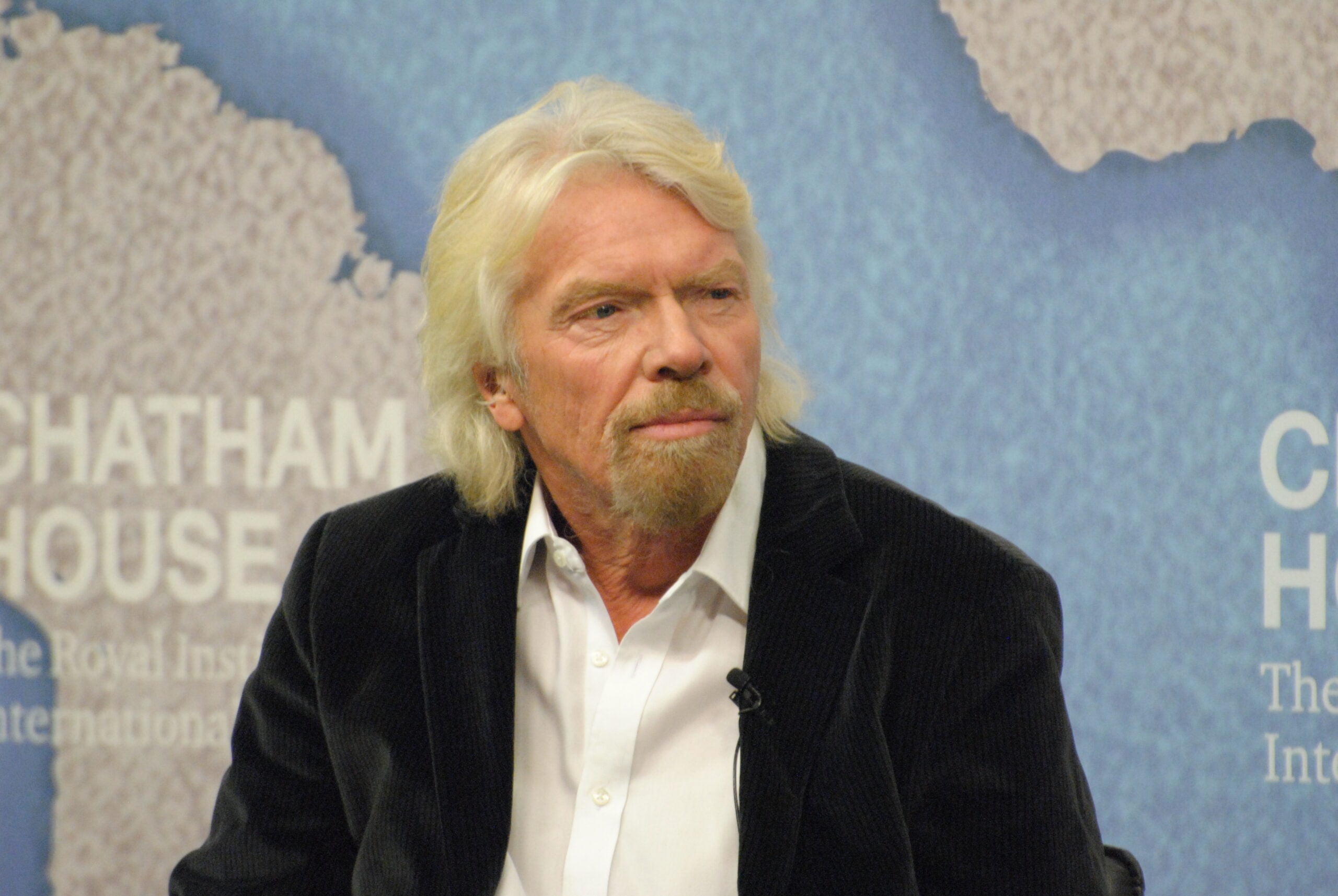 A man with long white hair and a beard, wearing a white shirt and dark jacket, sits in front of a blue background with "Chatham House" text partially visible.