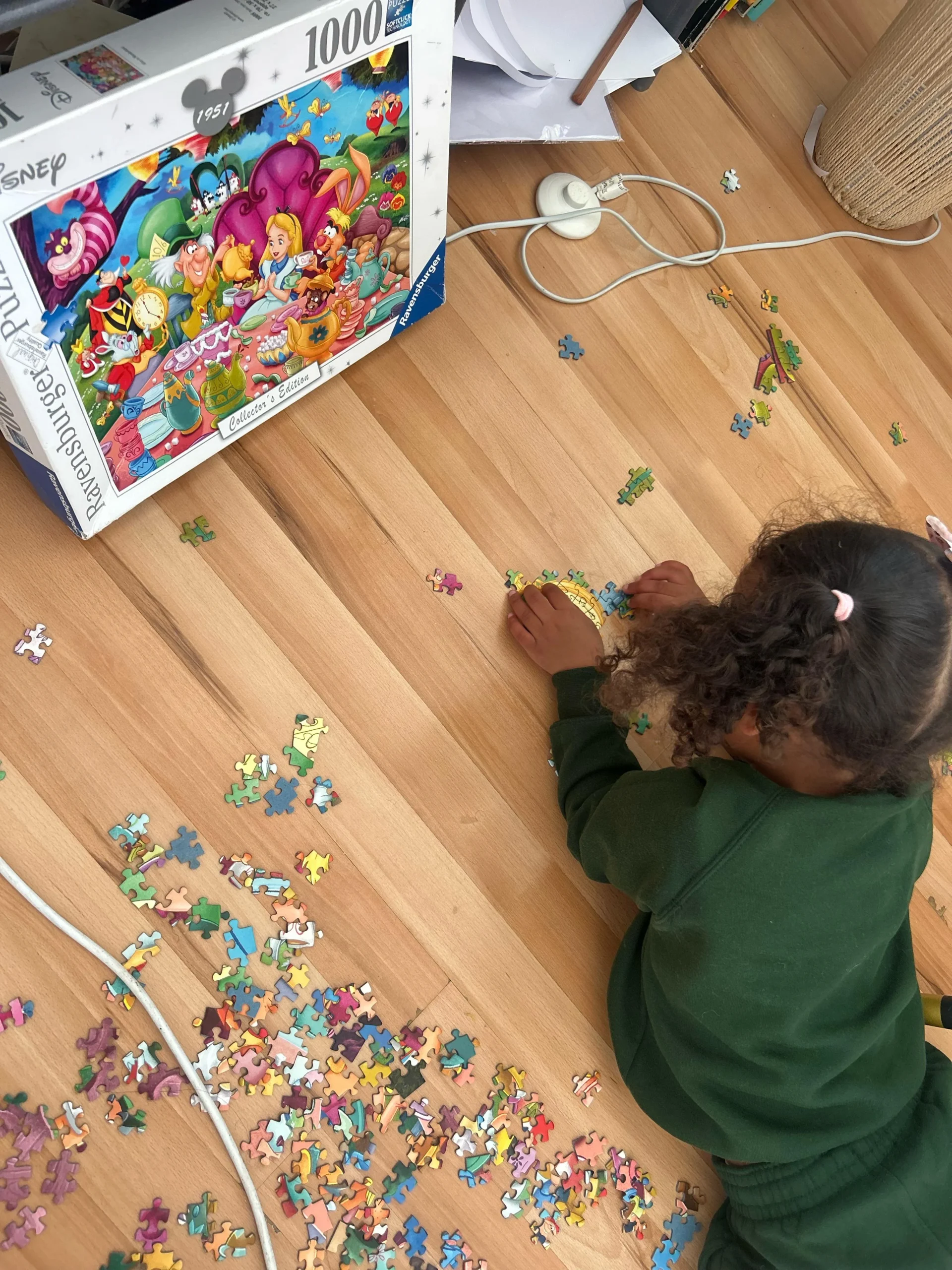 A young child in green clothes lies on a wooden floor, assembling a colorful Disney jigsaw puzzle. Puzzle pieces are scattered around, and the puzzle box with cartoon characters is propped up nearby. Electric cords are visible on the floor.