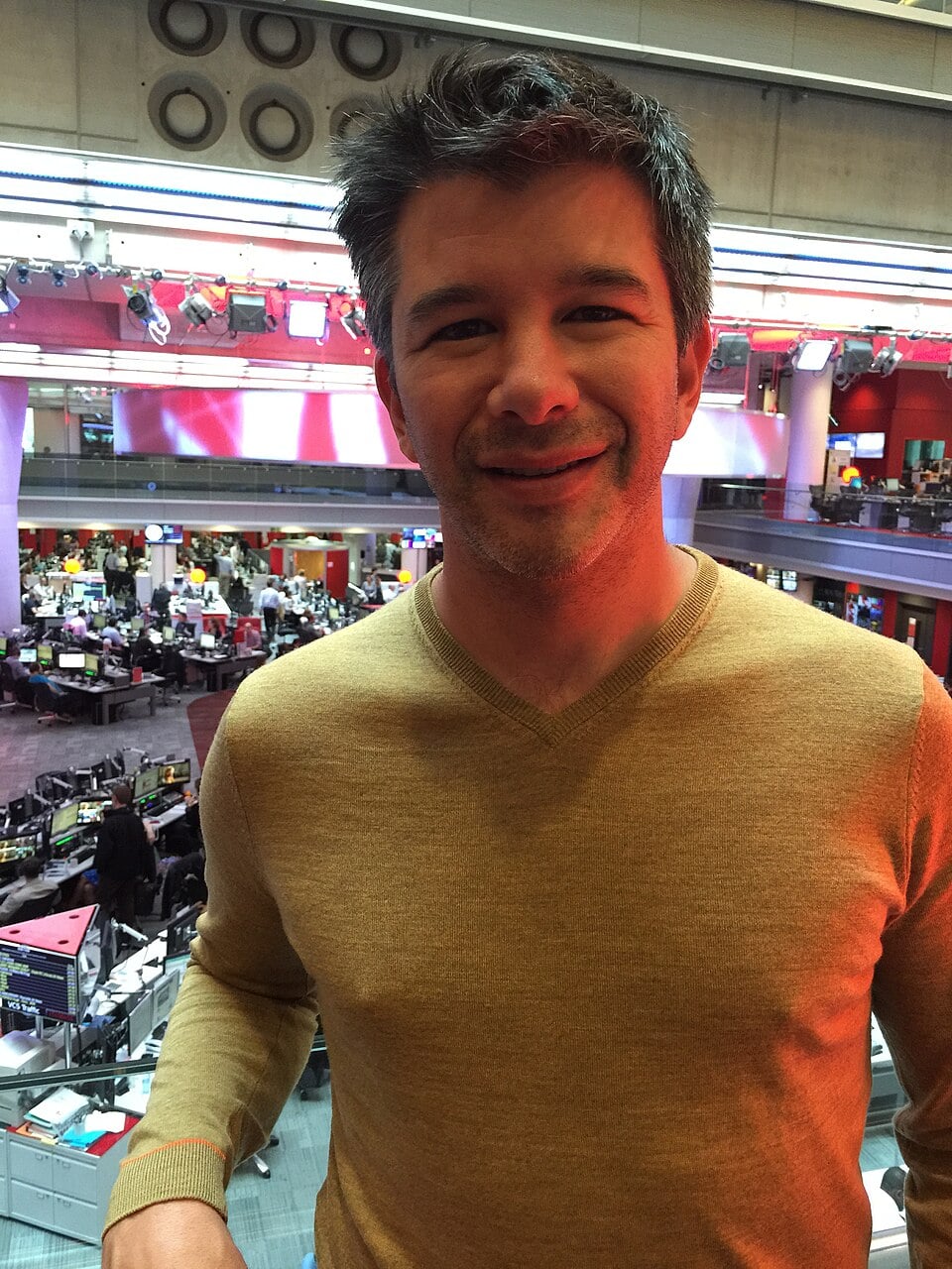 A man with short, dark hair wearing a light brown sweater is smiling and standing indoors with a busy newsroom and office area visible in the background.