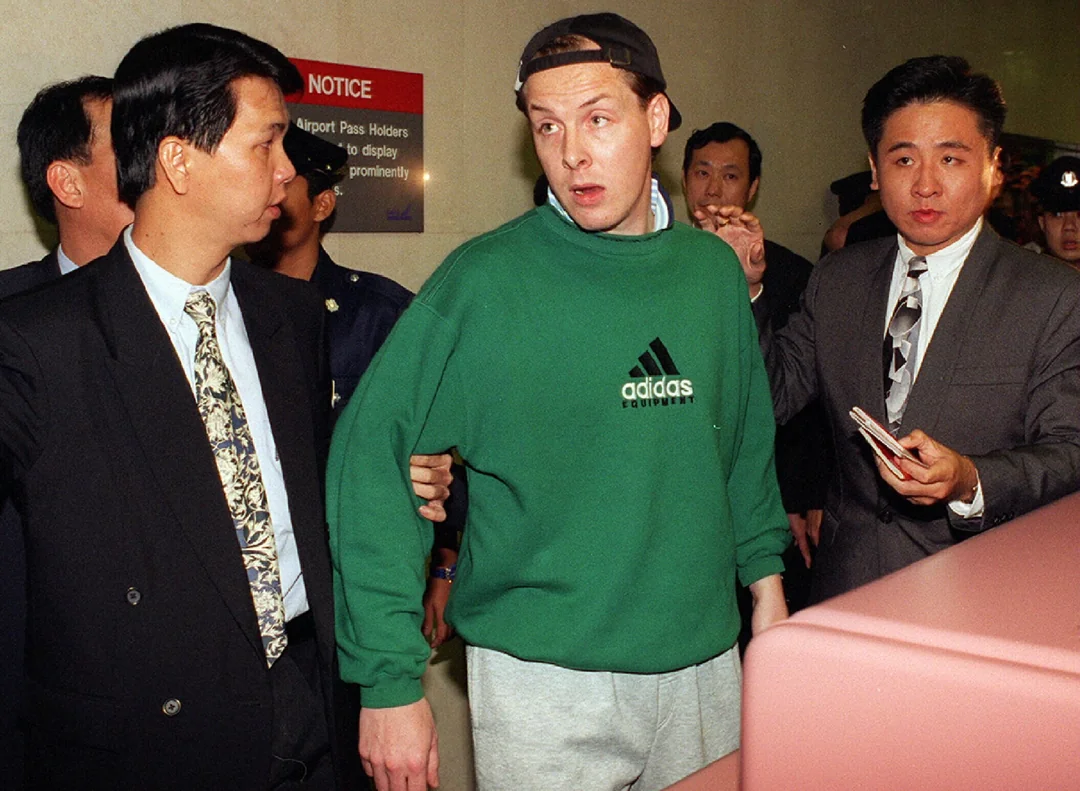 A man in a green Adidas sweatshirt is being escorted by several men in suits through a building. The man looks surprised or alarmed, while one suited man holds his arm and another gestures ahead.