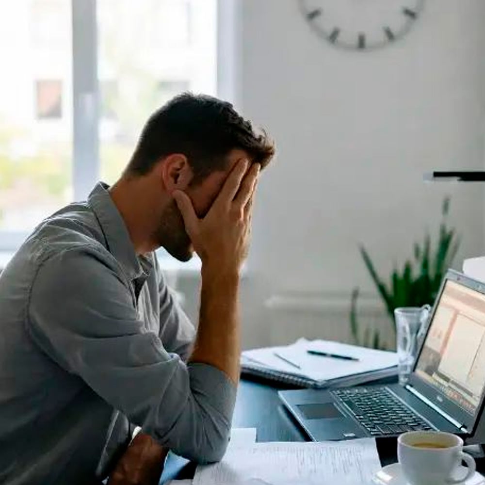 A man sits at a desk with his hand covering his face, looking stressed while working on a laptop. Papers, a cup, and a glass sit nearby; a clock and a plant are visible in the background.