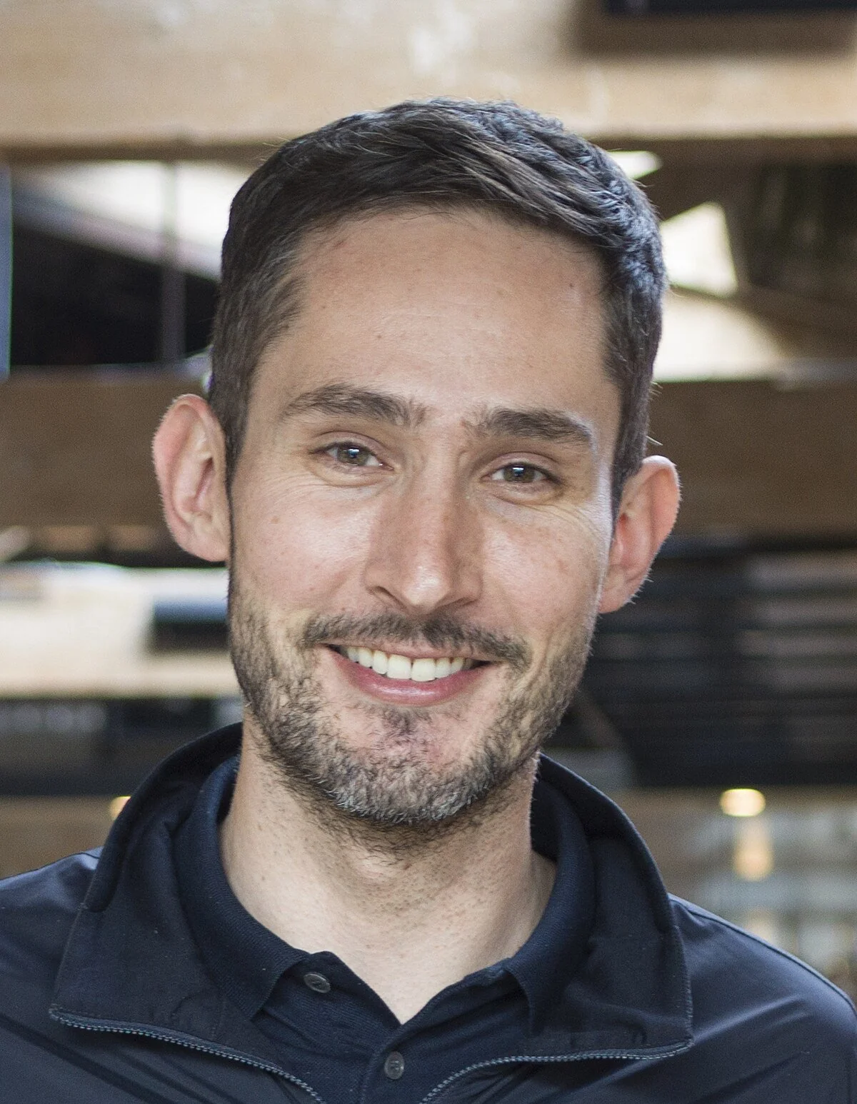 A man with short dark hair and a trimmed beard is smiling at the camera, wearing a black jacket and a dark shirt, standing indoors with blurred wooden beams and lights in the background.
