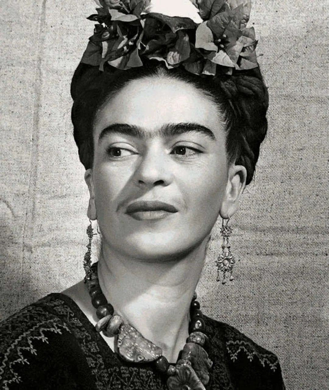 Black-and-white portrait of a woman with dark braided hair adorned with flowers, wearing large earrings, a beaded necklace, and a patterned top, posing in front of a plain fabric background.