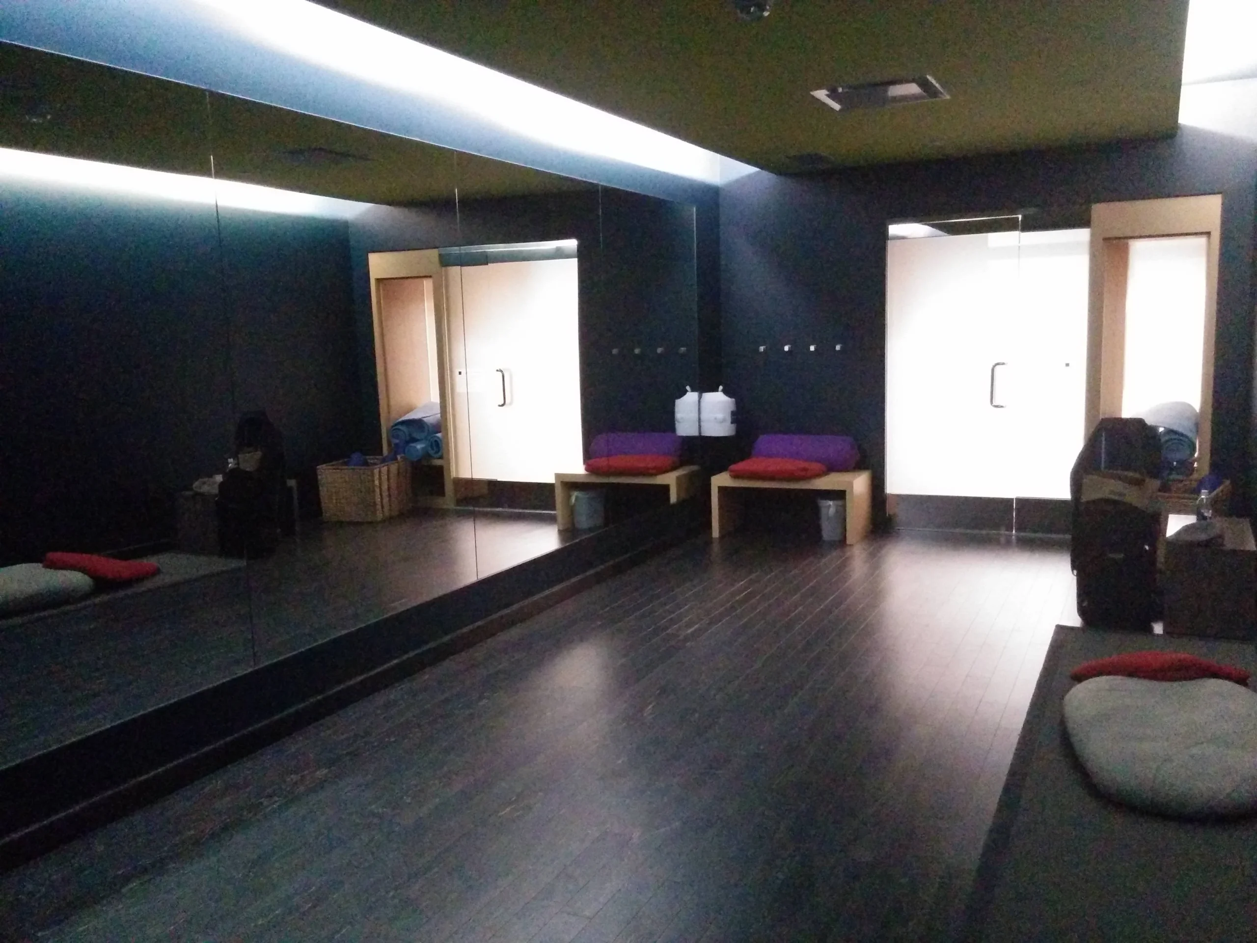 A dimly lit yoga or meditation room with dark floors, mirrored walls, cushioned benches, pillows, and baskets. Light streams in from two doors with frosted glass panels at the far end.