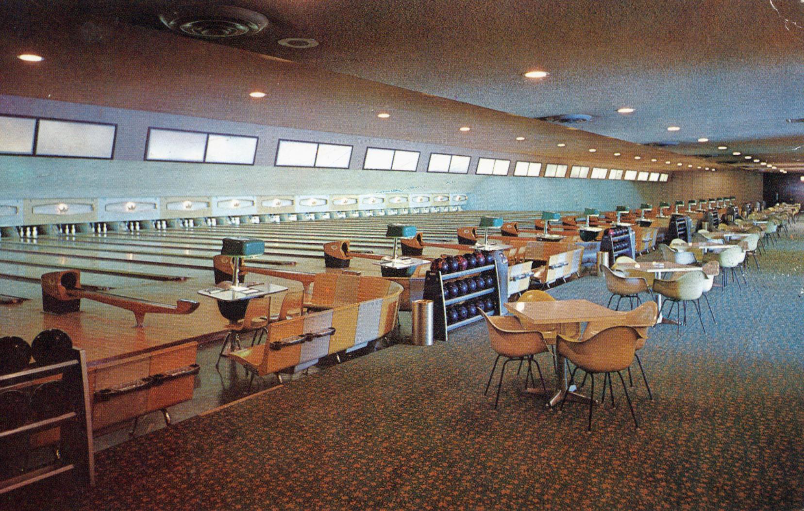 A vintage bowling alley with multiple lanes, retro seating, ball racks, and tables with chairs. The space is well-lit with ceiling lights and small windows, creating a mid-century modern atmosphere.