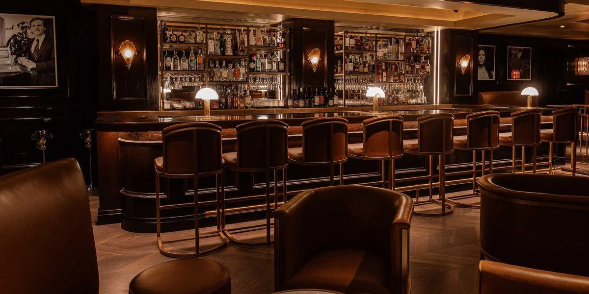 A dimly lit, elegant bar with brown leather chairs lined up at a long counter, shelves of liquor bottles behind the bar, and framed black-and-white photos on dark walls. Warm, ambient lighting creates a cozy atmosphere.