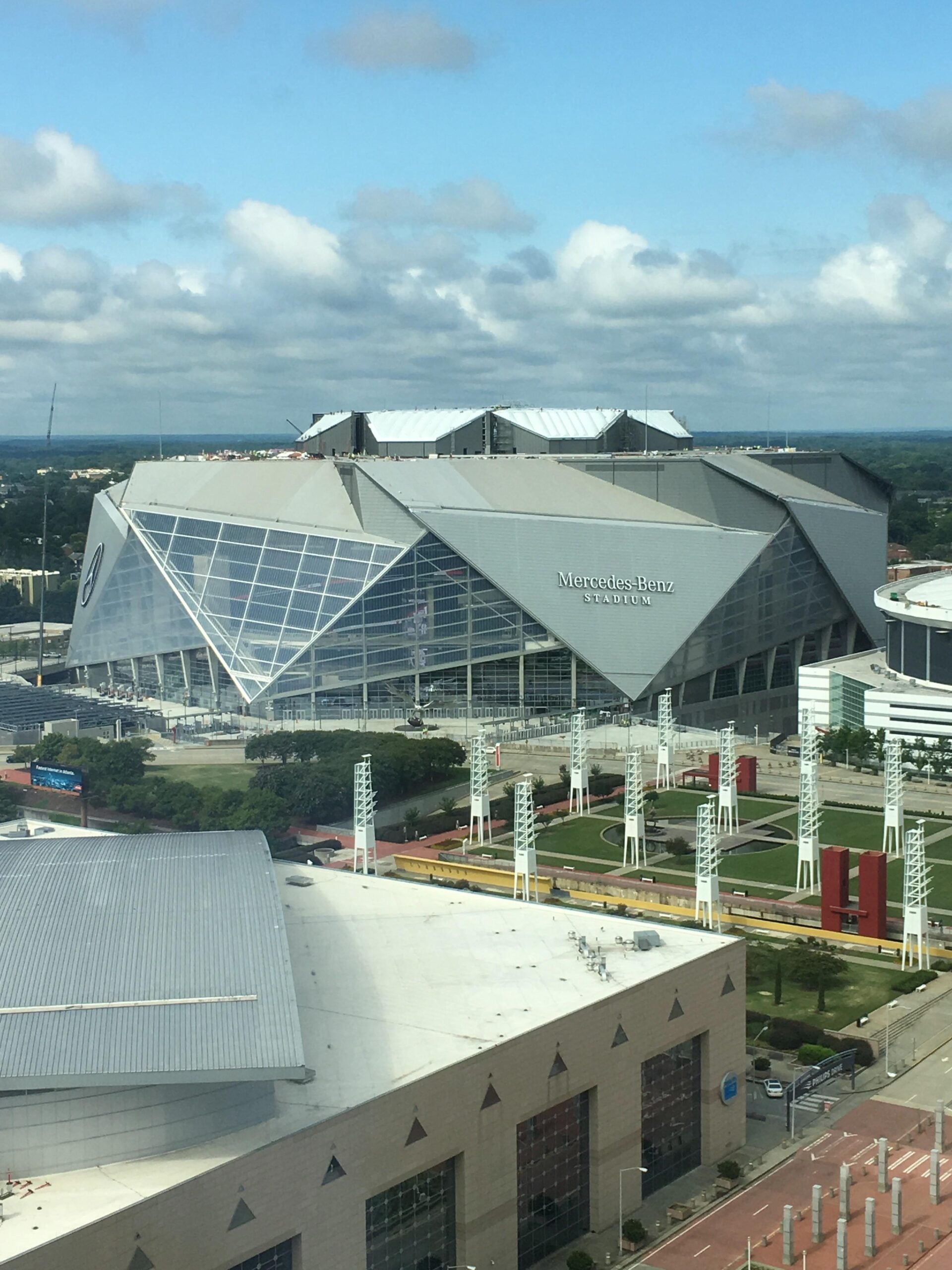 Aerial view of Mercedes-Benz Stadium in Atlanta, Georgia, featuring its distinctive angular roof design and large glass windows under a partly cloudy sky. Surrounding buildings and streets are visible.