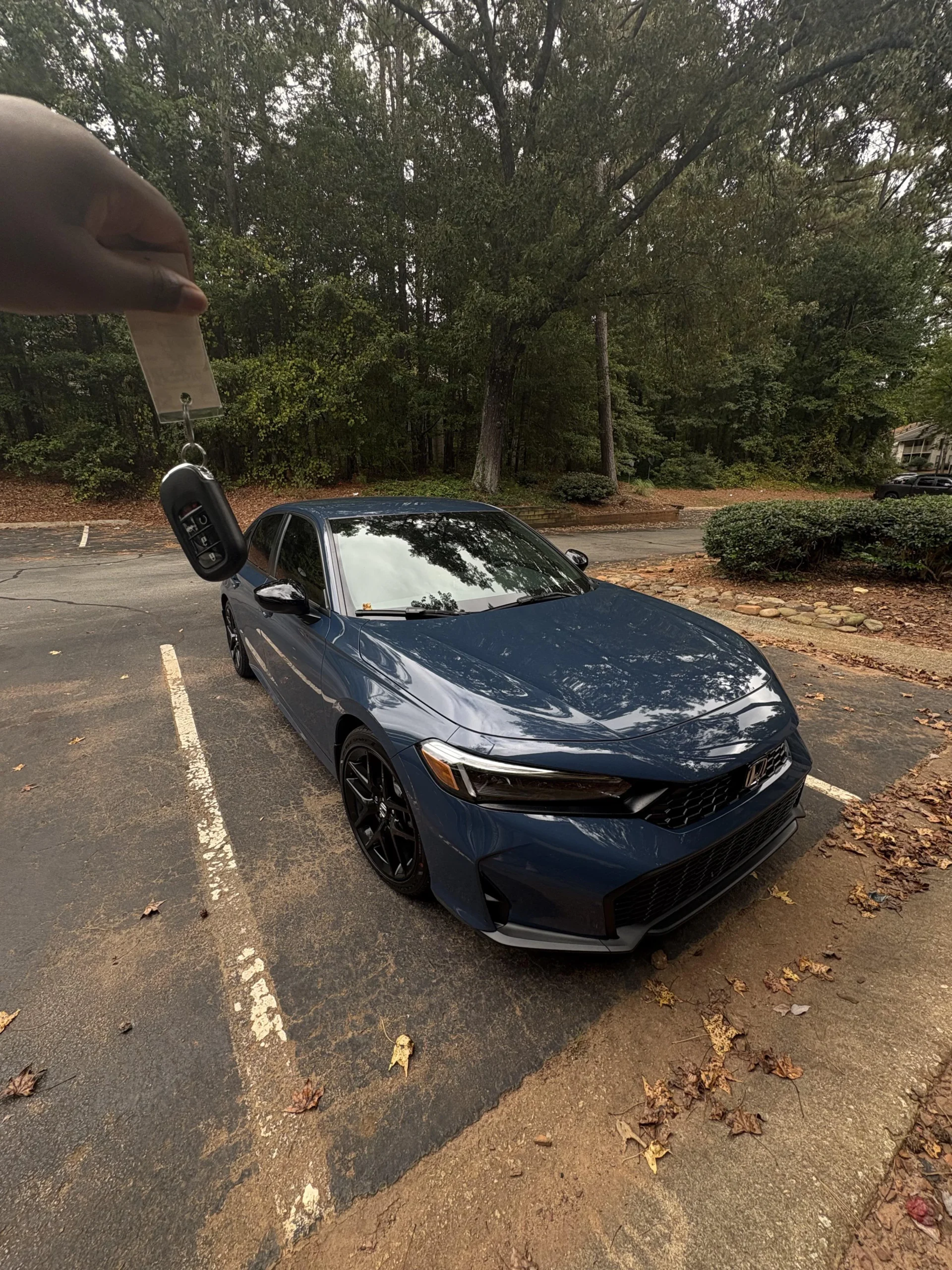 A person holds a car key fob in front of a blue sedan parked in a wooded, outdoor parking lot on an overcast day. Fallen leaves are scattered on the ground around the car.