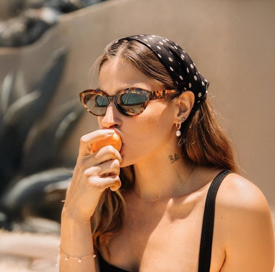 A woman wearing cat-eye sunglasses and a black polka-dot headscarf eats a peach. She has long hair, a black top, earrings, and a necklace, enjoying a sunny outdoor setting with blurred plants in the background.