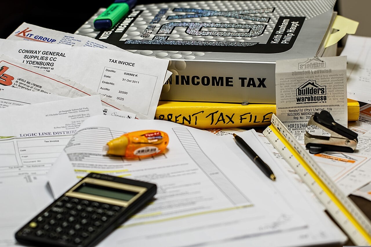 A cluttered desk with tax documents, invoices, a calculator, pen, highlighter, correction tape, and thick folders labeled "INCOME TAX" and "CURRENT TAX FILES," representing tax preparation and financial paperwork.