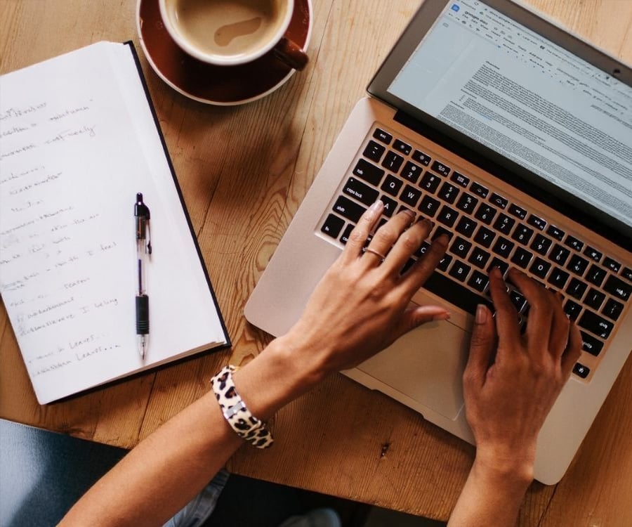 A person types on a laptop at a wooden table with a cup of coffee, a notebook filled with handwritten notes, and a pen nearby.