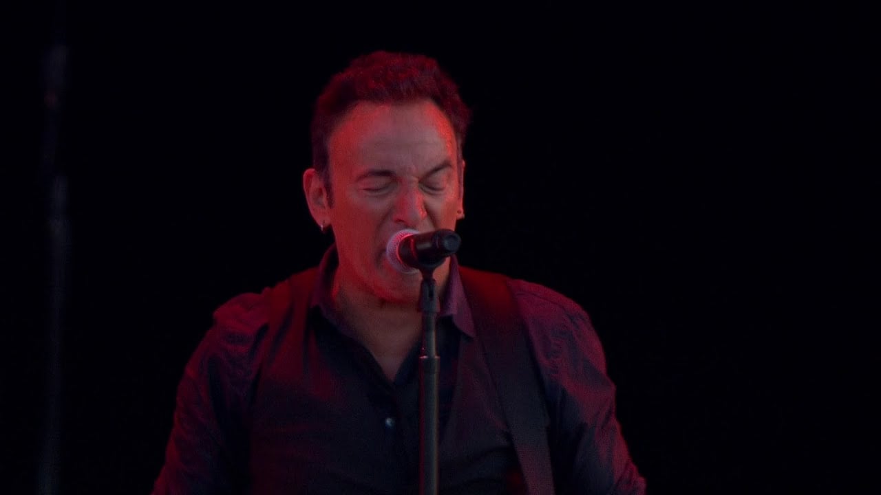 A person with short dark hair sings passionately into a microphone on stage, wearing a dark buttoned shirt, against a black background.