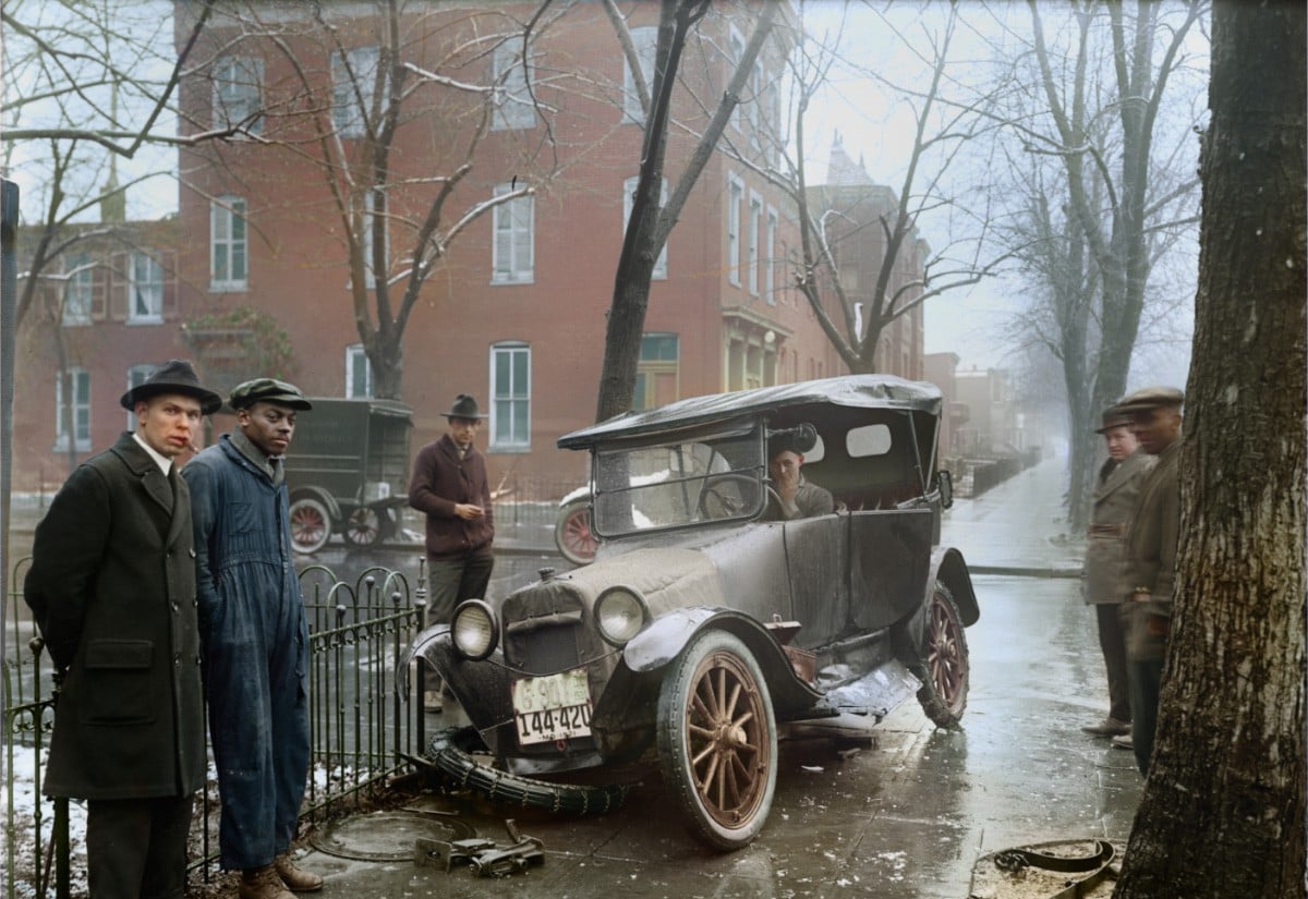 A group of men stand around an old car with a damaged wheel on a wet, snowy city street lined with bare trees and brick buildings. One man sits in the car while others watch.