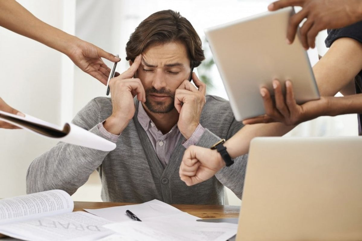 A man sits at a cluttered desk, eyes closed and fingers on his temples, looking stressed as multiple hands around him offer documents, a tablet, a pen, and show a watch, suggesting he is overwhelmed at work.