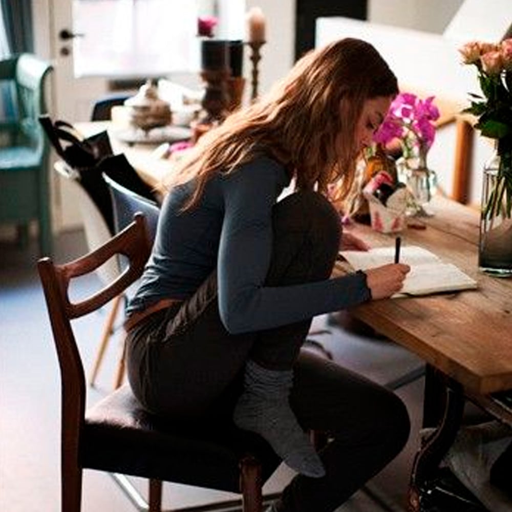 A woman with long hair sits cross-legged on a chair at a wooden table, writing in a notebook. The room is softly lit, with flowers and candles on the table and various household items in the background.