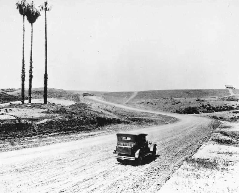 A vintage car drives on a winding dirt road through a barren landscape with three tall palm trees on the left and a small building in the distance under a clear sky.