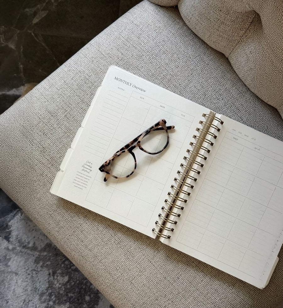 An open planner displaying a monthly overview page rests on a light textured chair, with a pair of tortoiseshell eyeglasses placed on top of the planner.