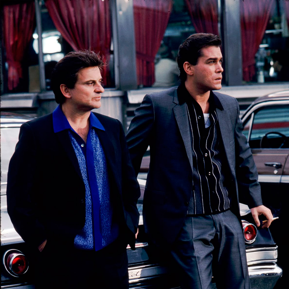 Two men in suits stand in front of a classic car outside a diner with red curtains, both looking off into the distance with serious expressions.