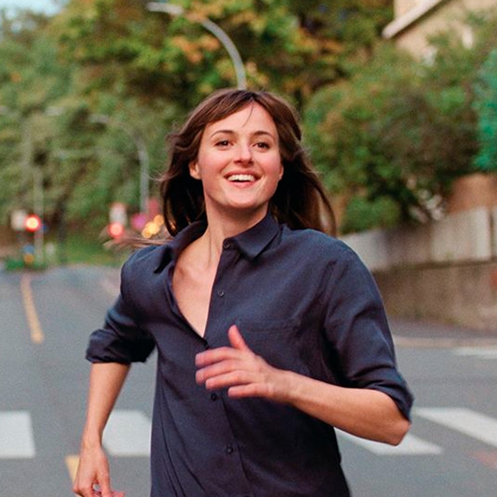 A woman with brown hair, wearing a dark blue shirt, is running and smiling on a street lined with trees and buildings. The background is slightly blurred, giving a sense of movement.