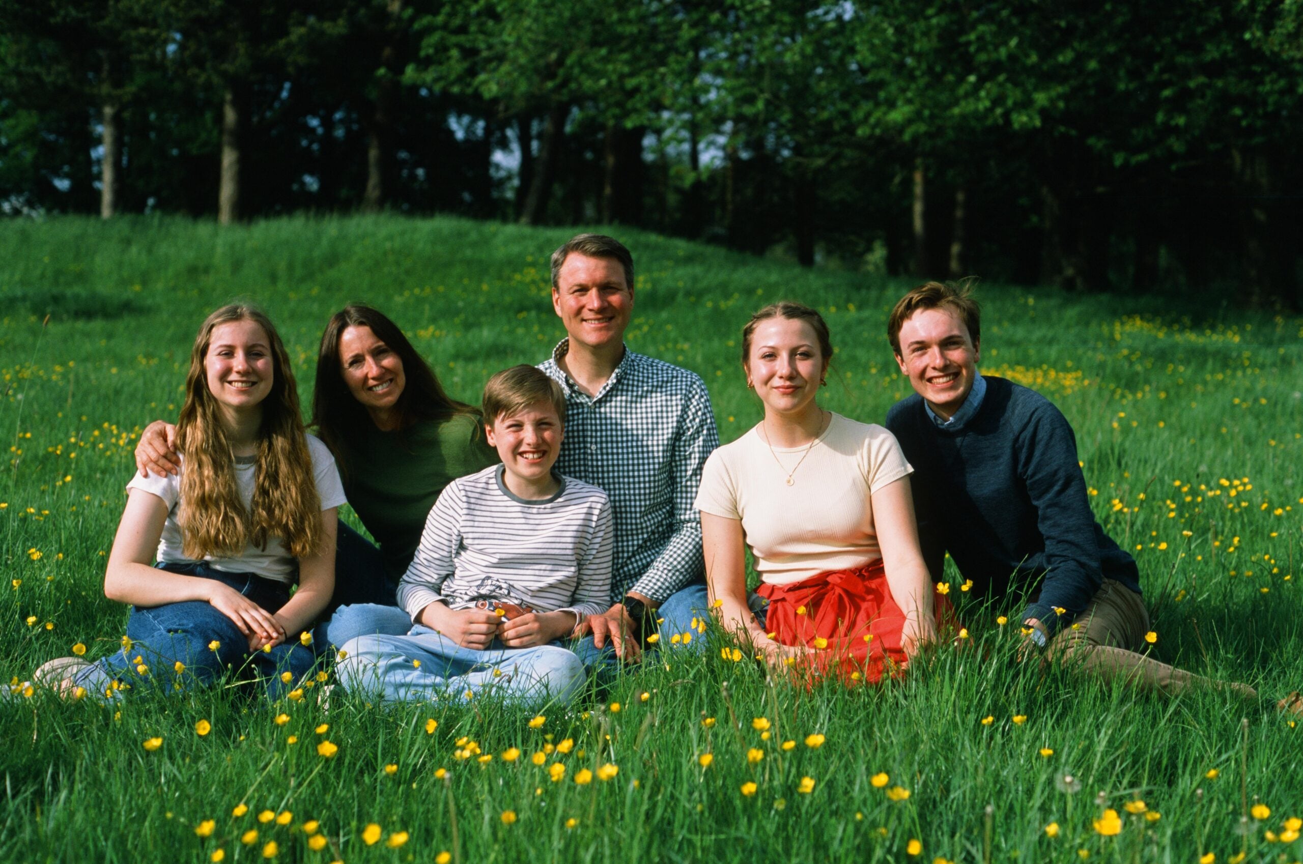Six people sit closely together on green grass with yellow wildflowers, smiling at the camera. Trees and sunlight fill the background, creating a warm and cheerful outdoor scene.