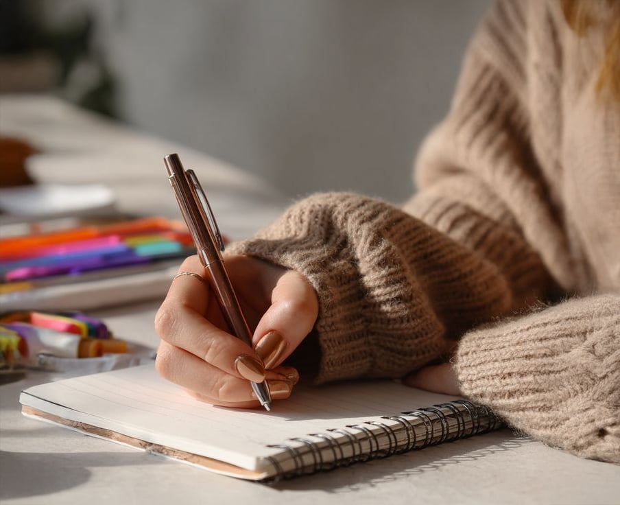 A person wearing a cozy brown sweater is writing in a spiral notebook with a pen. Colorful markers and pens are scattered on the table in the background.