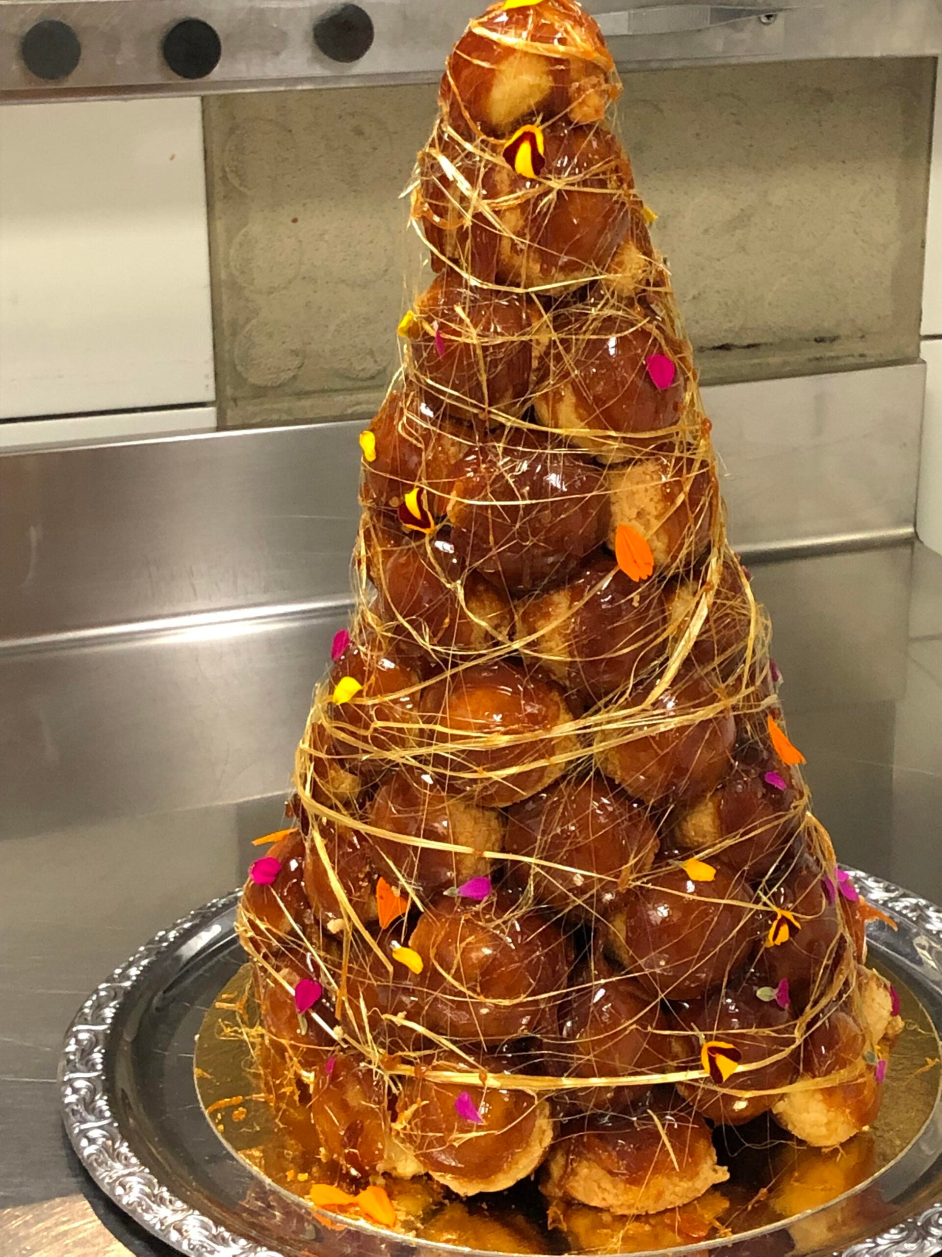 A tall croquembouche tower made of caramel-coated cream puffs, decorated with spun sugar and colorful edible flower petals, sits on a silver platter in a kitchen setting.