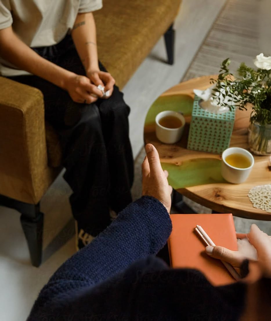 Two people sit across from each other in a cozy setting, one holding a notebook and pen, the other with hands clasped. A table between them holds tissues, cups of tea, and a vase of flowers, suggesting a therapy session.