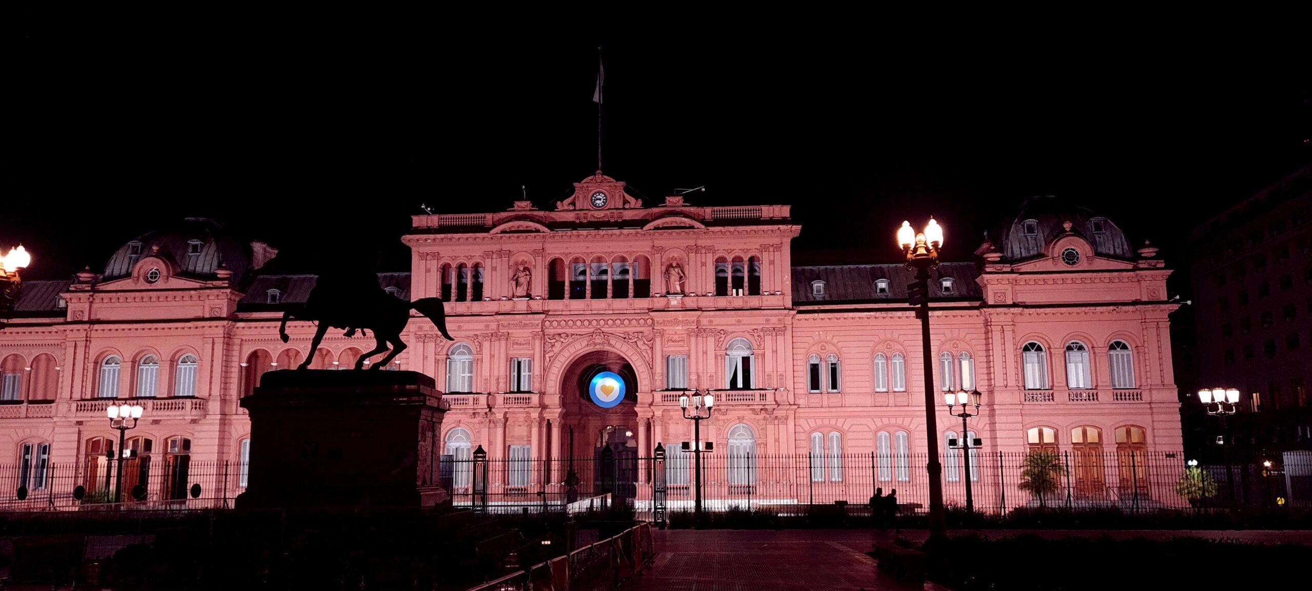 A large pink government building, Casa Rosada, is illuminated at night. A statue of a person on horseback is silhouetted in the foreground, and street lamps light the plaza.