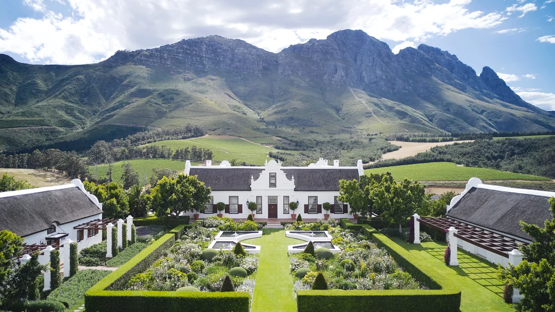 A large, white Cape Dutch-style manor with thatched roofs sits in a manicured garden, surrounded by green lawns and hedges, with dramatic mountains rising in the background under a partly cloudy sky.