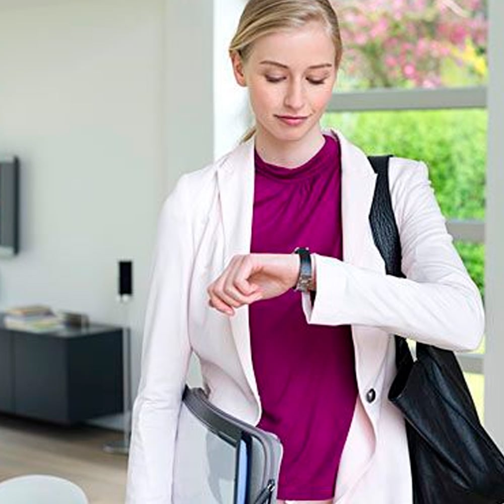 A woman in a white suit and magenta top checks her smartwatch while holding a laptop and a black shoulder bag, standing indoors near a window with greenery outside.