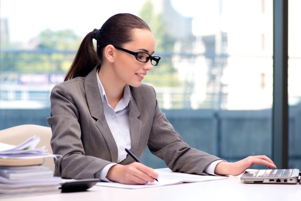 A woman in a gray suit and glasses sits at a desk, writing on paper with one hand and using a laptop with the other. There are stacks of documents on the desk and large windows in the background.