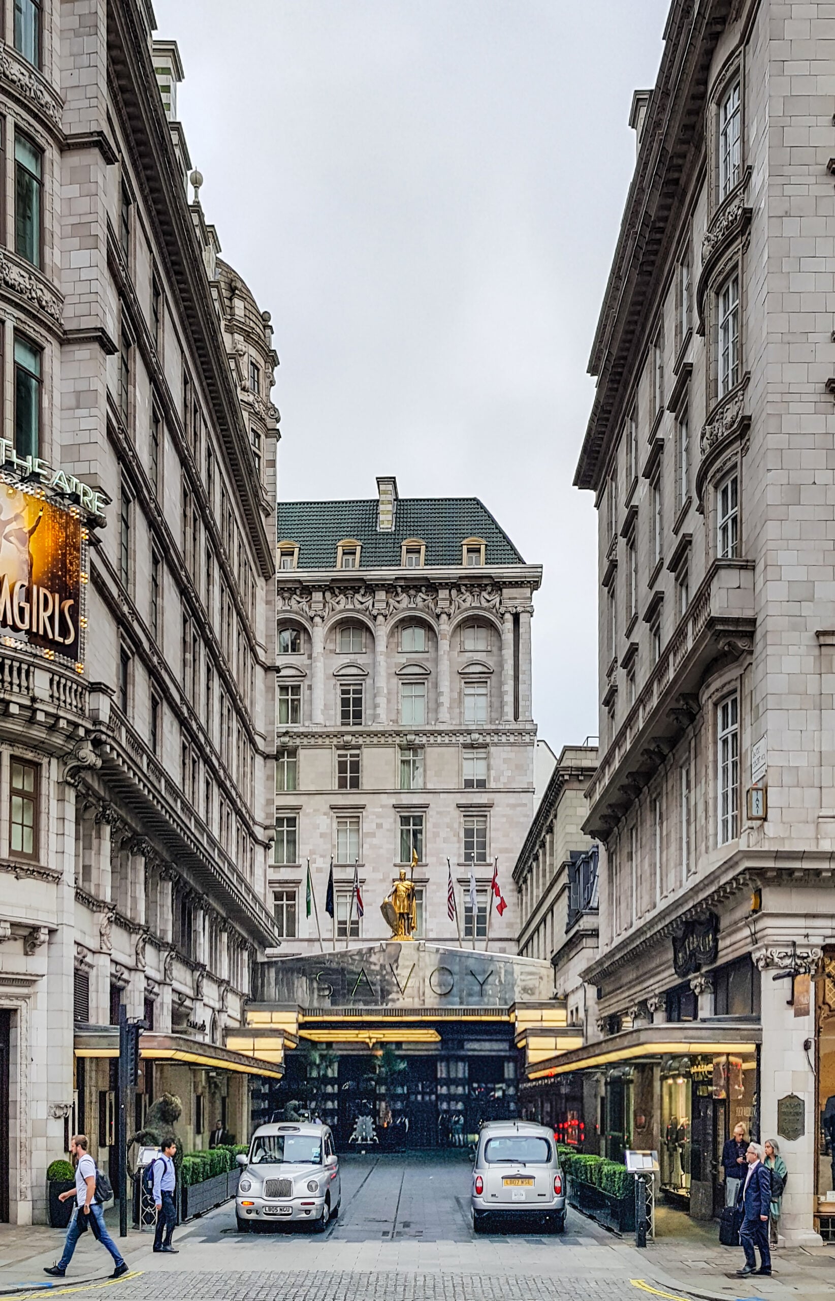 A grand hotel entrance with flags, two cars parked out front, and people walking nearby, flanked by tall, ornate historic buildings on a city street.
