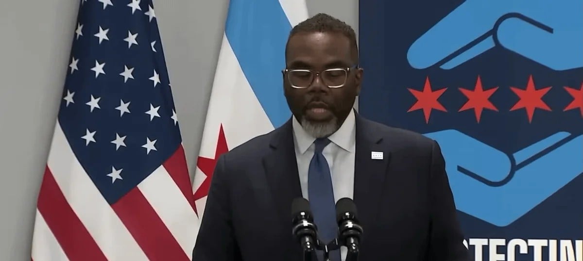 A man in a suit and glasses speaks at a podium with microphones, standing in front of the U.S. flag, the Chicago flag, and a blue backdrop featuring red stars and blue hands.
