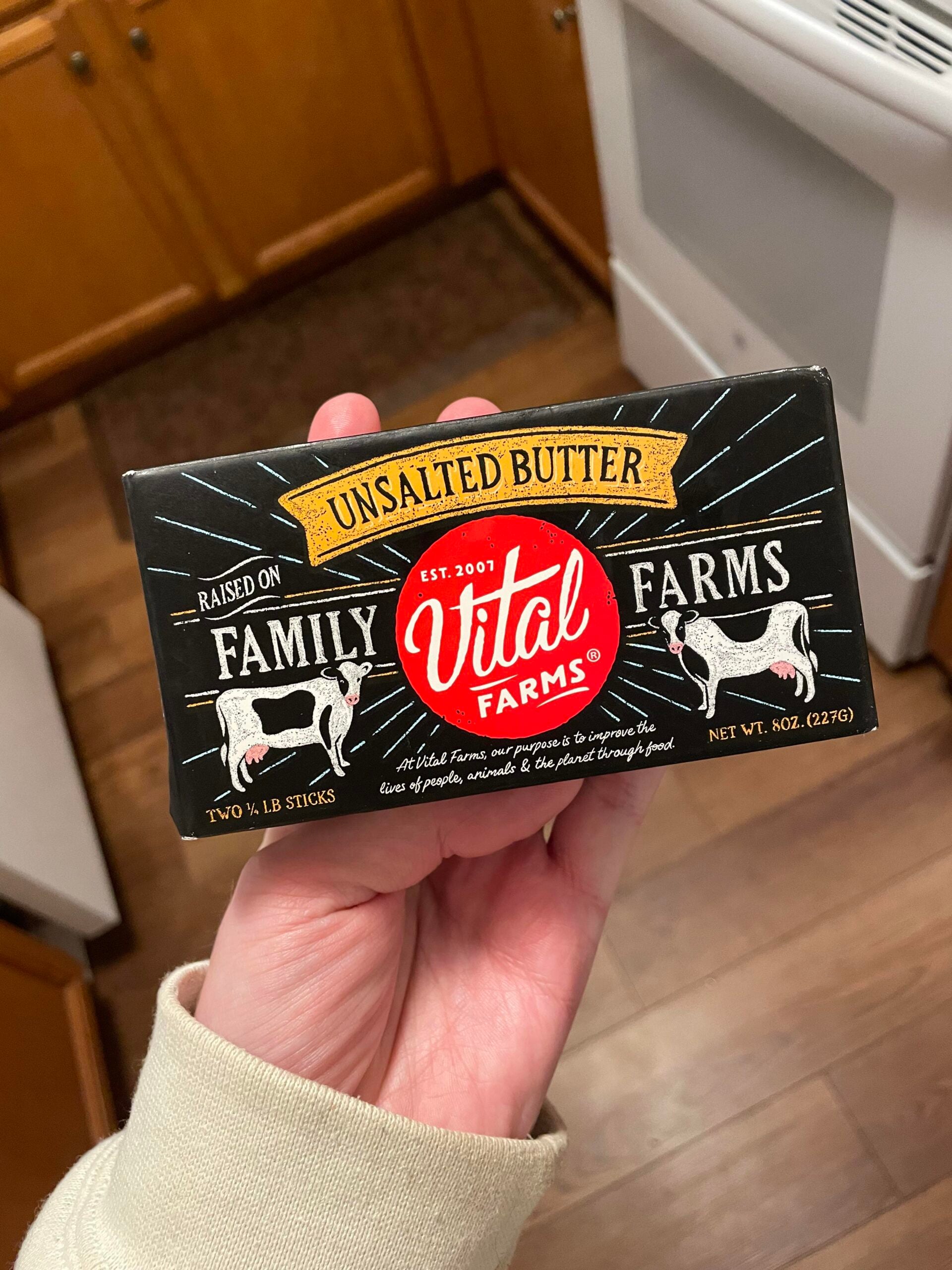 A hand holds a package of Vital Farms unsalted butter, featuring black packaging with white and red text, cow illustrations, and kitchen cabinets visible in the background.