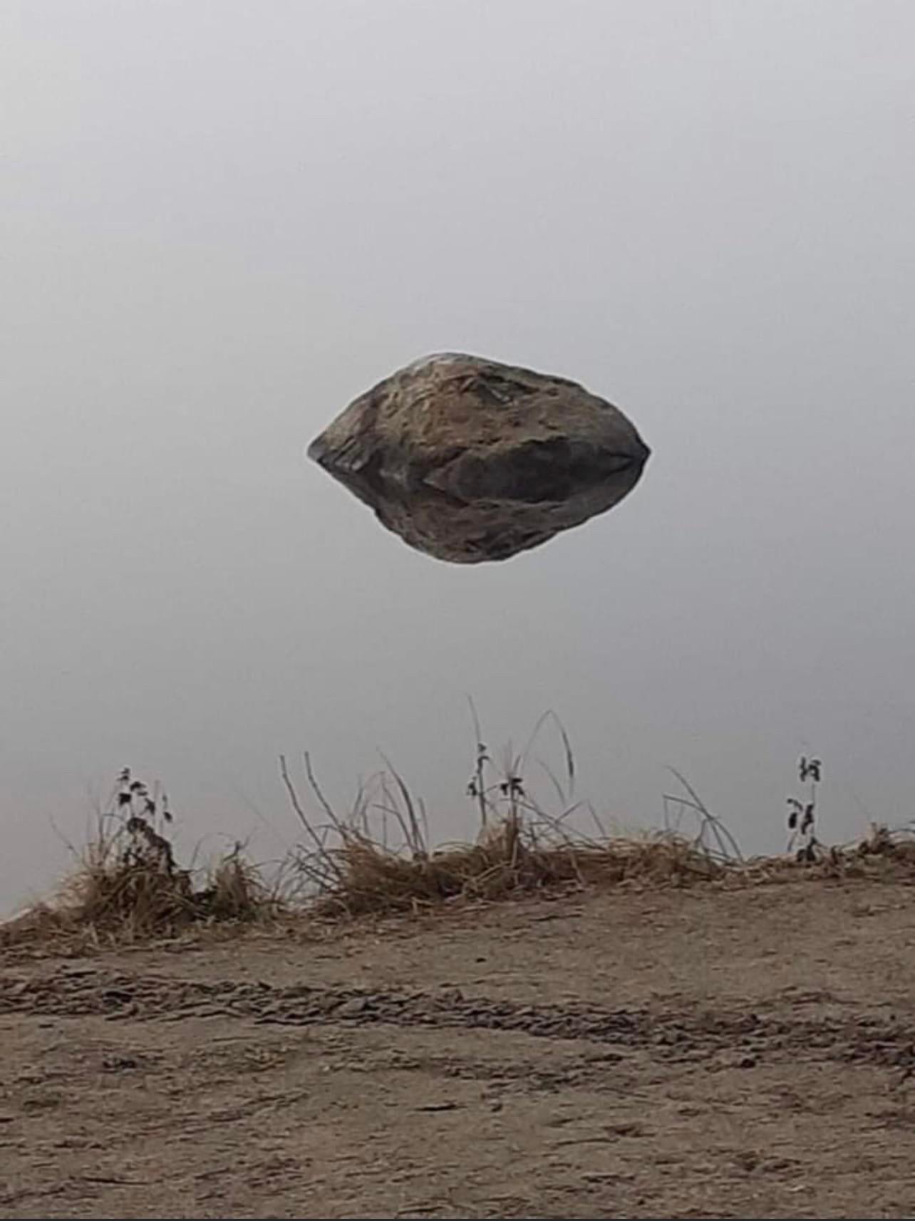 A large rock appears to be floating in midair above a calm, misty body of water, with dry grass and dirt in the foreground. The rock's reflection in the water creates the illusion of levitation.