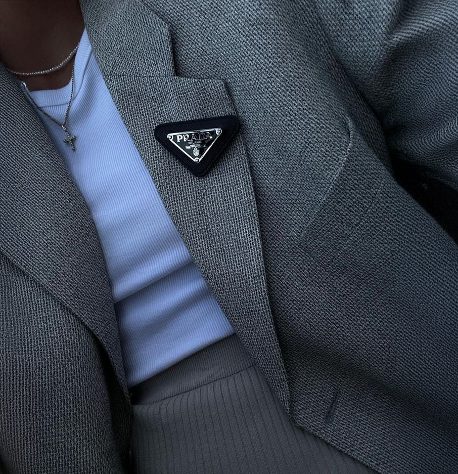 A person wearing a light gray textured blazer over a white top, accessorized with a thin chain necklace featuring a cross pendant and a triangular Prada logo pin on the blazer lapel.