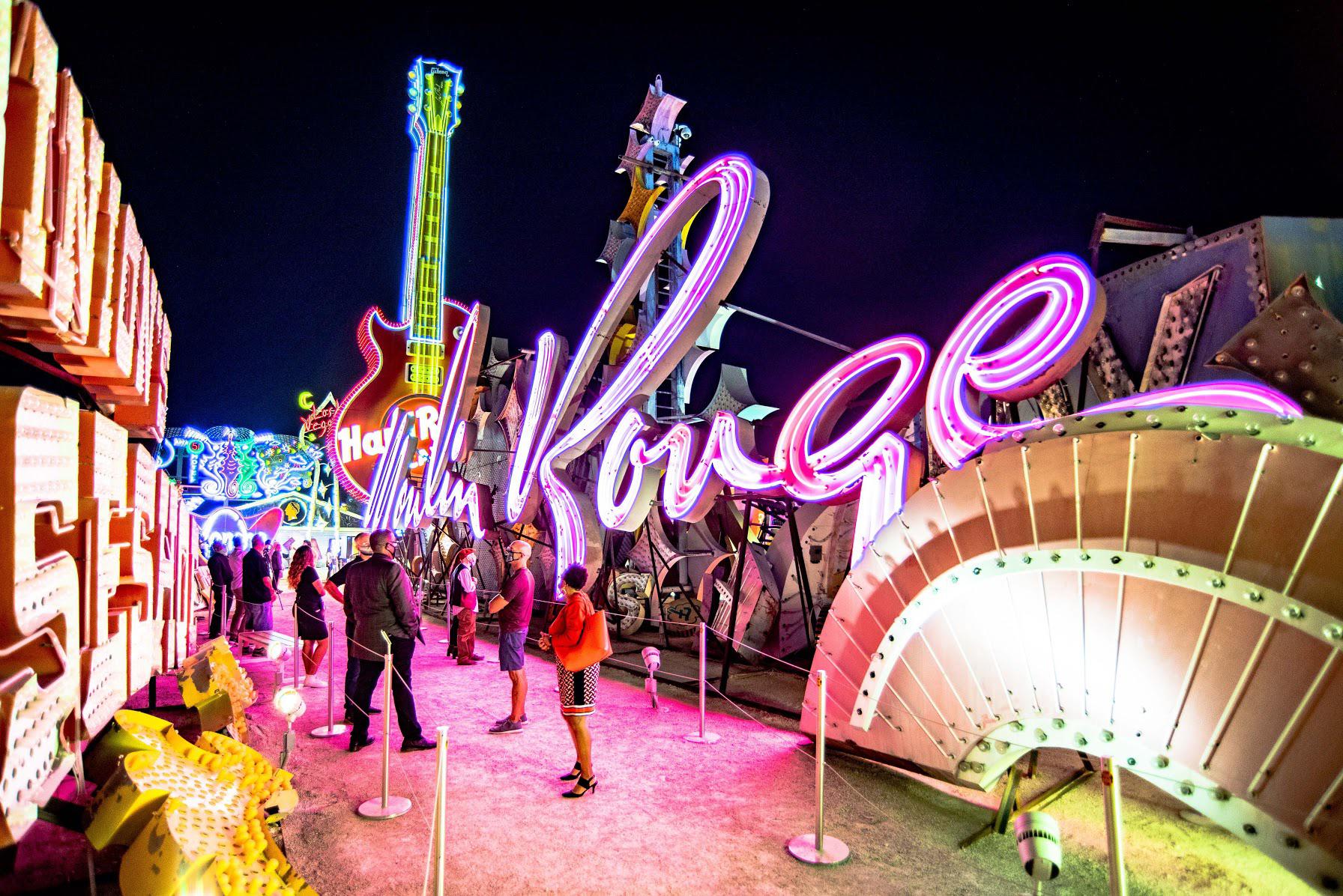 People walk along a path lined with colorful, glowing vintage neon signs at night, including a large pink Moulin Rouge sign and a red Hard Rock Cafe guitar. The scene is lively and vibrant.