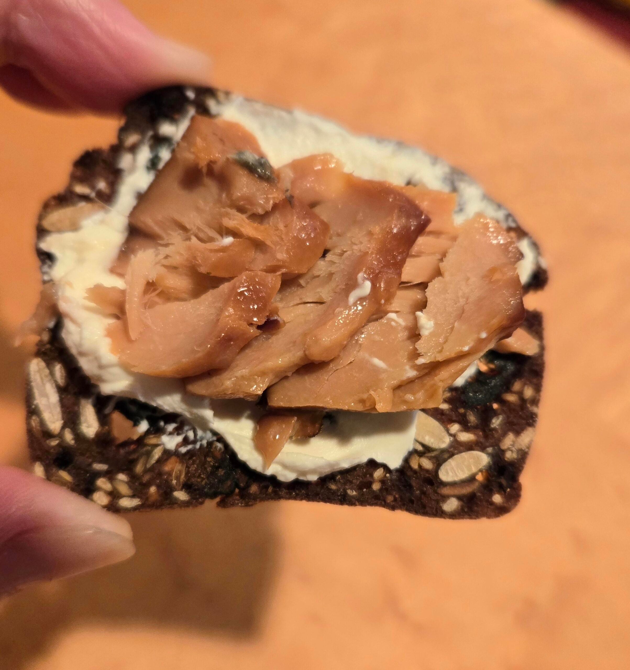 A hand holds a seeded cracker topped with a layer of creamy white cheese and chunks of flaked smoked salmon, against an orange background.