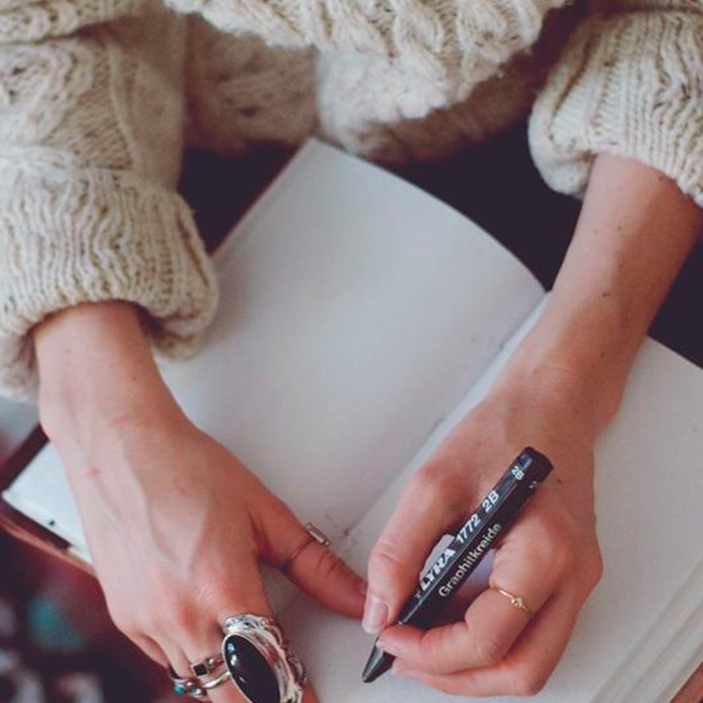 A person wearing a chunky knit sweater writes in a blank notebook with a black pen, adorned with rings on their fingers. The scene suggests journaling or note-taking in a cozy setting.