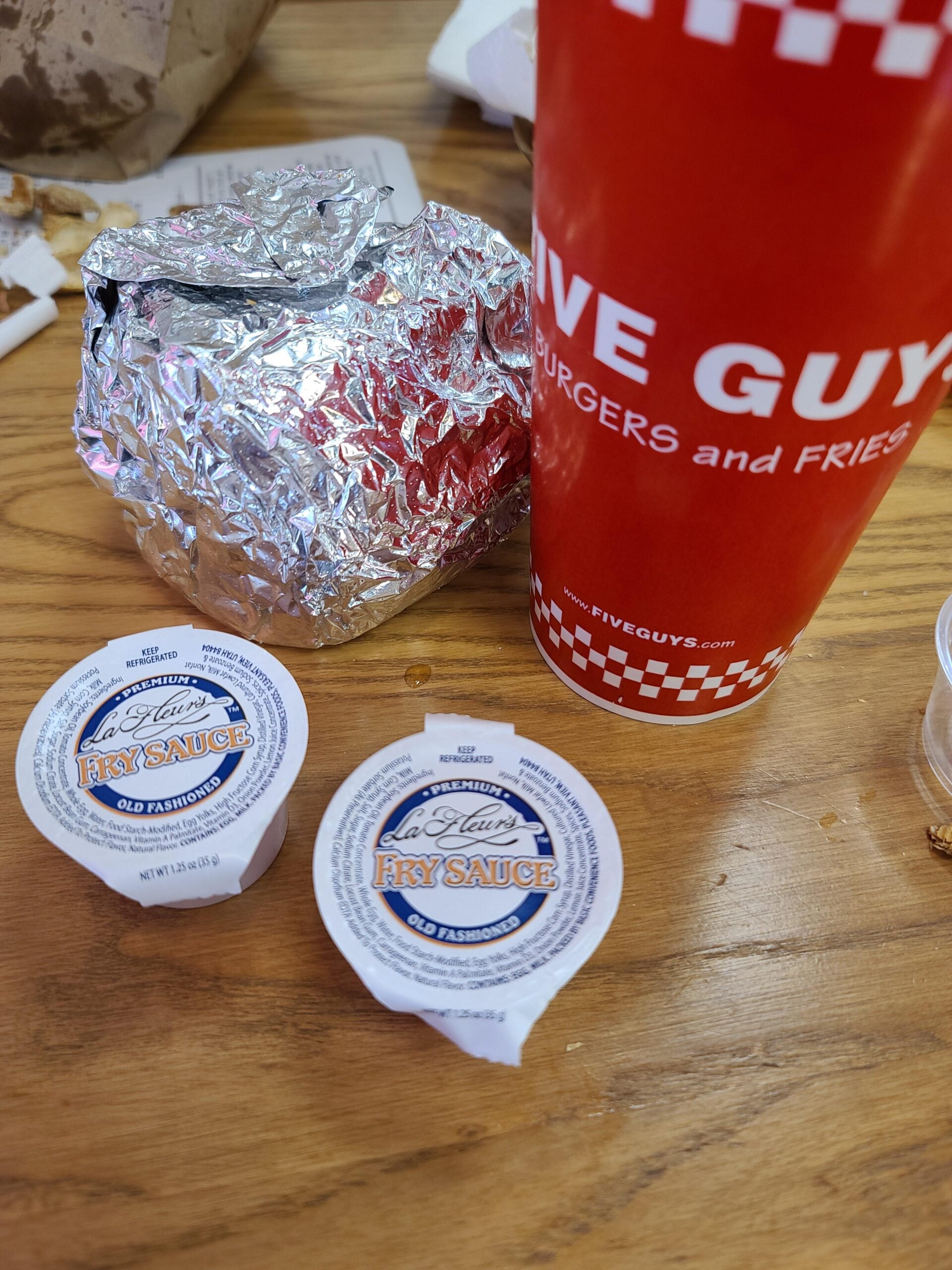 A wrapped burger, a large Five Guys drink cup, and two tubs of La Beau’s Fry Sauce sit on a wooden table. Some food wrappers and crumbs are visible in the background.