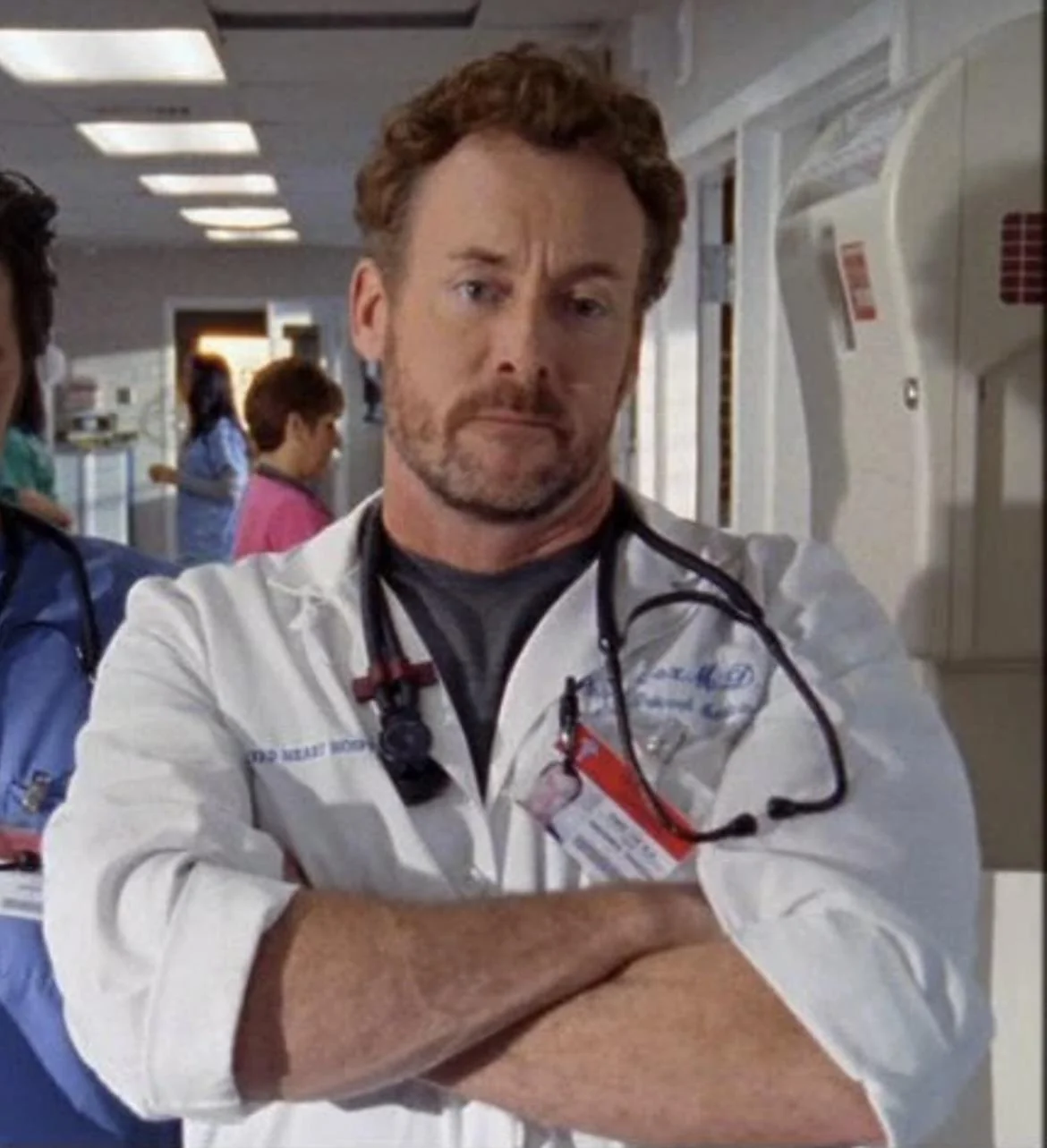 A male doctor with a beard and curly hair stands in a hospital corridor with his arms crossed, wearing a white coat and stethoscope. Other medical staff are visible in the background.