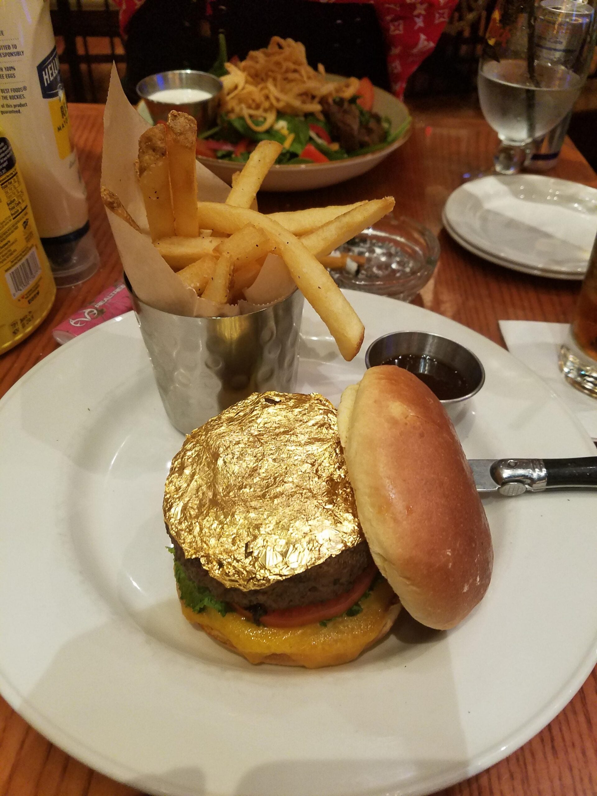 A hamburger topped with gold leaf sits on a bun next to a metal cup of French fries on a white plate. A small dish of sauce and a steak knife are also on the plate.