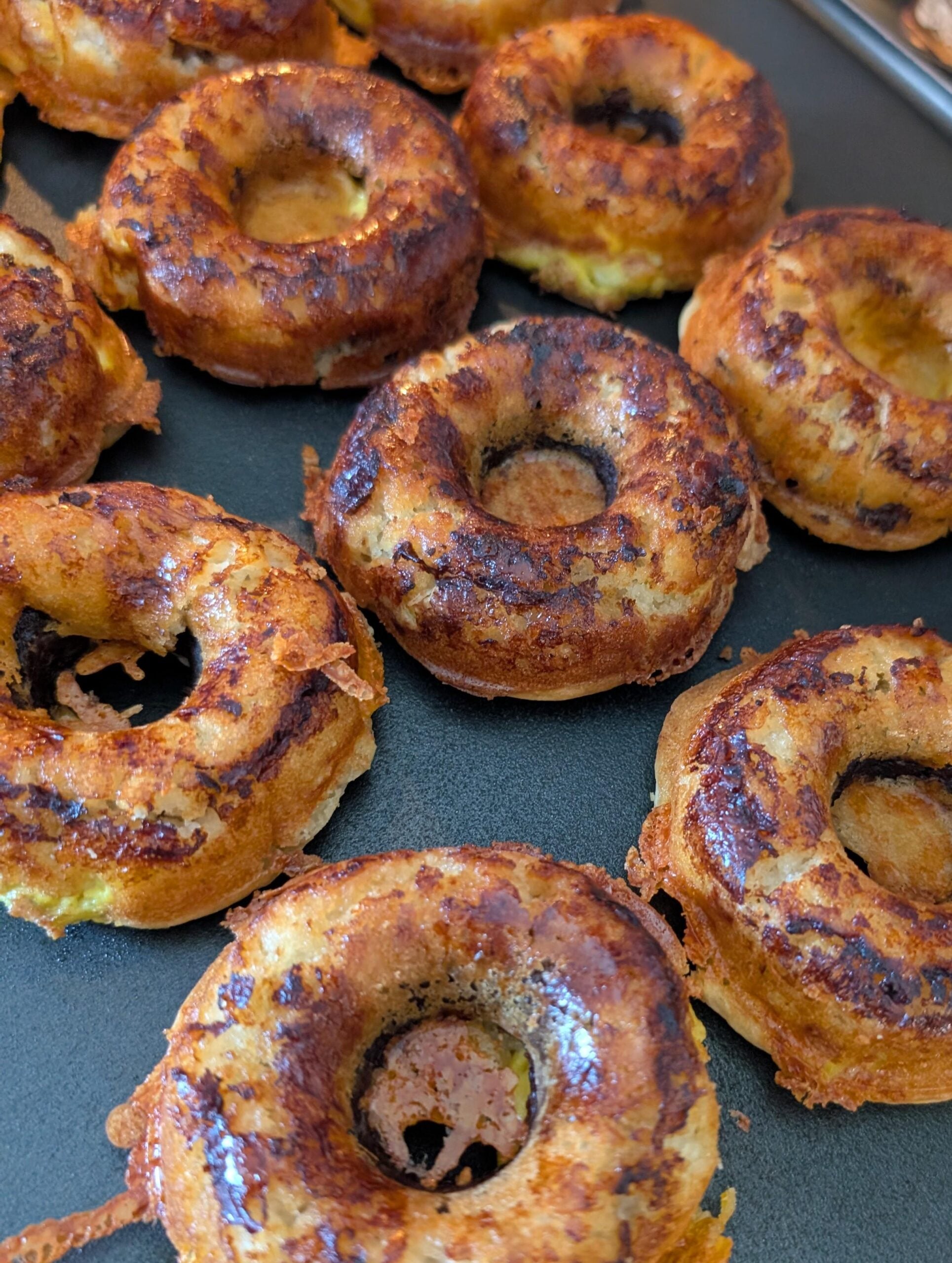 Golden brown, crispy, baked bagel-shaped items with a slightly uneven, rustic texture, arranged closely together on a dark baking tray. Some cheese appears to have melted and crisped around the edges.