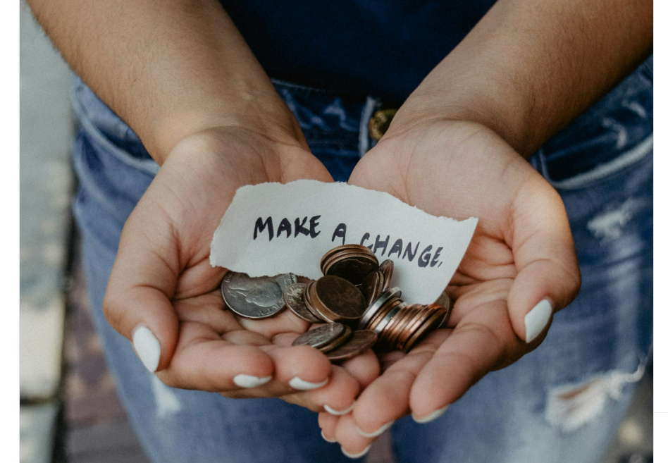 A person wearing jeans holds coins and a torn piece of paper with the handwritten message "MAKE A CHANGE" in their cupped hands.