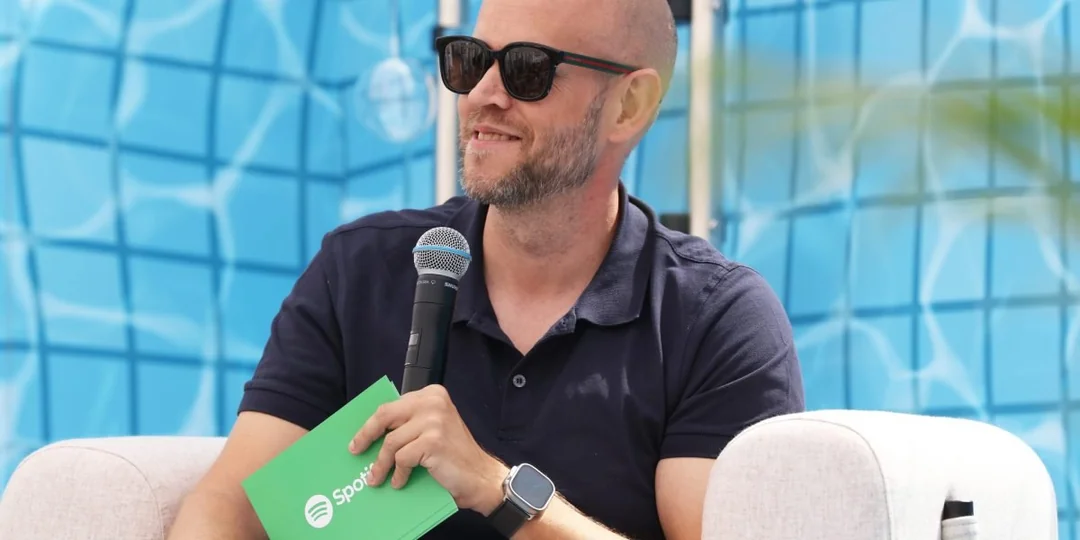 A man wearing sunglasses and a navy shirt sits on a light chair, holding a microphone and a green folder with the Spotify logo. Blue pool tiles are visible in the background.