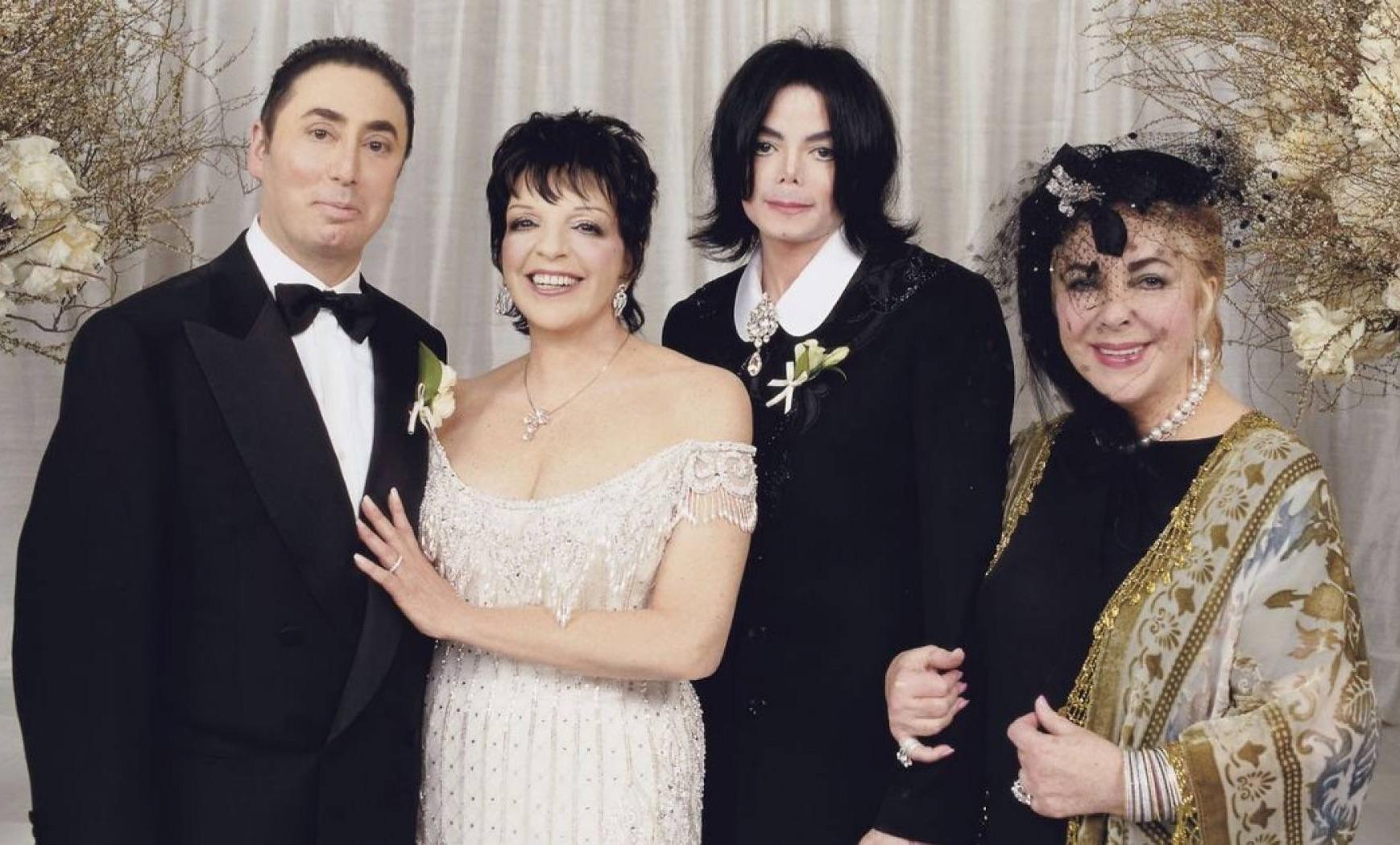 Four adults pose for a formal photo. Two men in tuxedos and two women in elegant dresses and accessories stand together, smiling, against a backdrop decorated with flowers and branches.