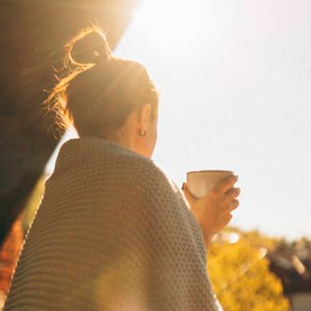 A person with hair in a bun, wrapped in a gray blanket, holds a mug while standing outside in bright sunlight, facing away from the camera toward a clear sky and trees.