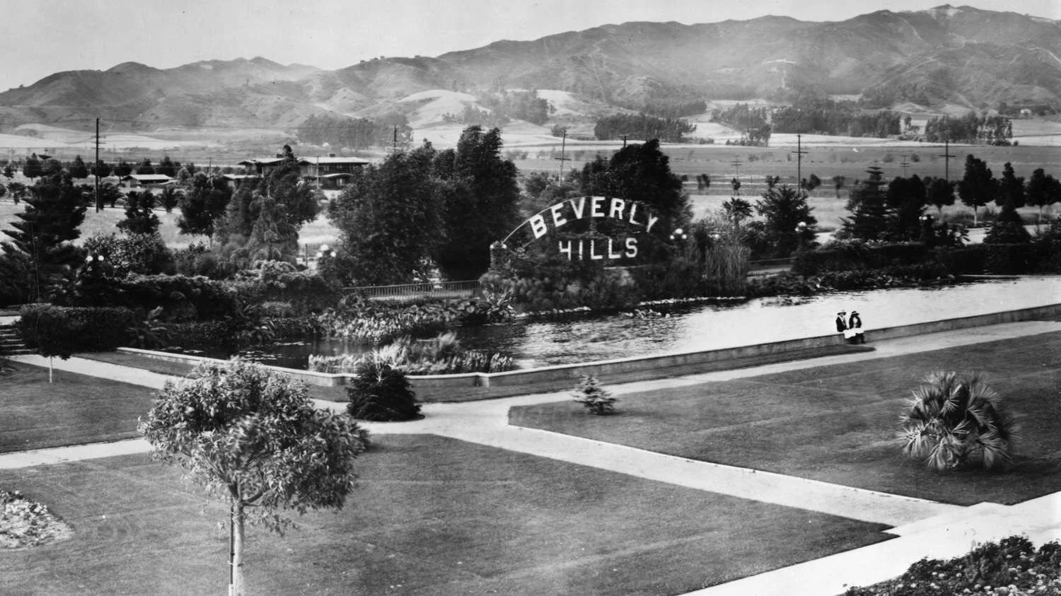 Black and white photo of a park with a pond and a large “Beverly Hills” sign. Two people sit on a bench by the water, with palm trees and mountains visible in the background.