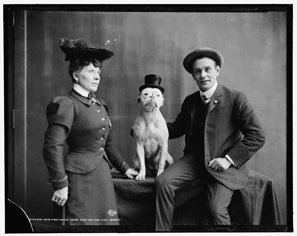 A woman and a man in vintage clothing pose for a portrait with a dog wearing a small top hat. The man is seated, the woman stands, and the dog sits between them on a covered table.