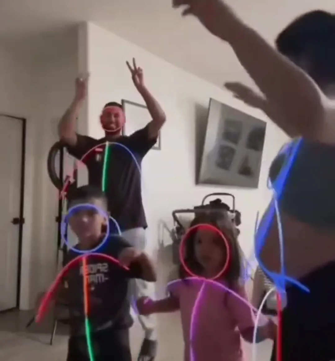 Four people, two adults and two children, are dancing indoors. Colorful neon lines outline their bodies and faces, creating stick figures. The adults have their arms raised, and everyone appears to be enjoying the activity.